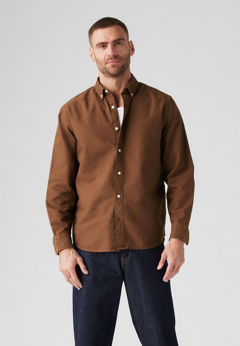 Camisa marrón de botones, fabricada en algodón, de corte relajado, con mangas largas y puños abotonados, combinada con jeans de mezclilla azul oscuro. Diseño sencillo, sin patrones.