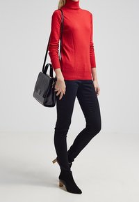 Roter Rollkragenpullover mit langen Ärmeln, kombiniert mit schwarzen Skinny Jeans und Ankle Boots. Eine schwarze Handtasche mit vertikalen Streifen rundet den Look ab.