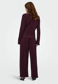Pruimkleurig, geplooid kostuum met een getailleerde blazer en wijde broekspijpen. De stof heeft verticale ribbels en strakke, minimalistische hardware-accenten.