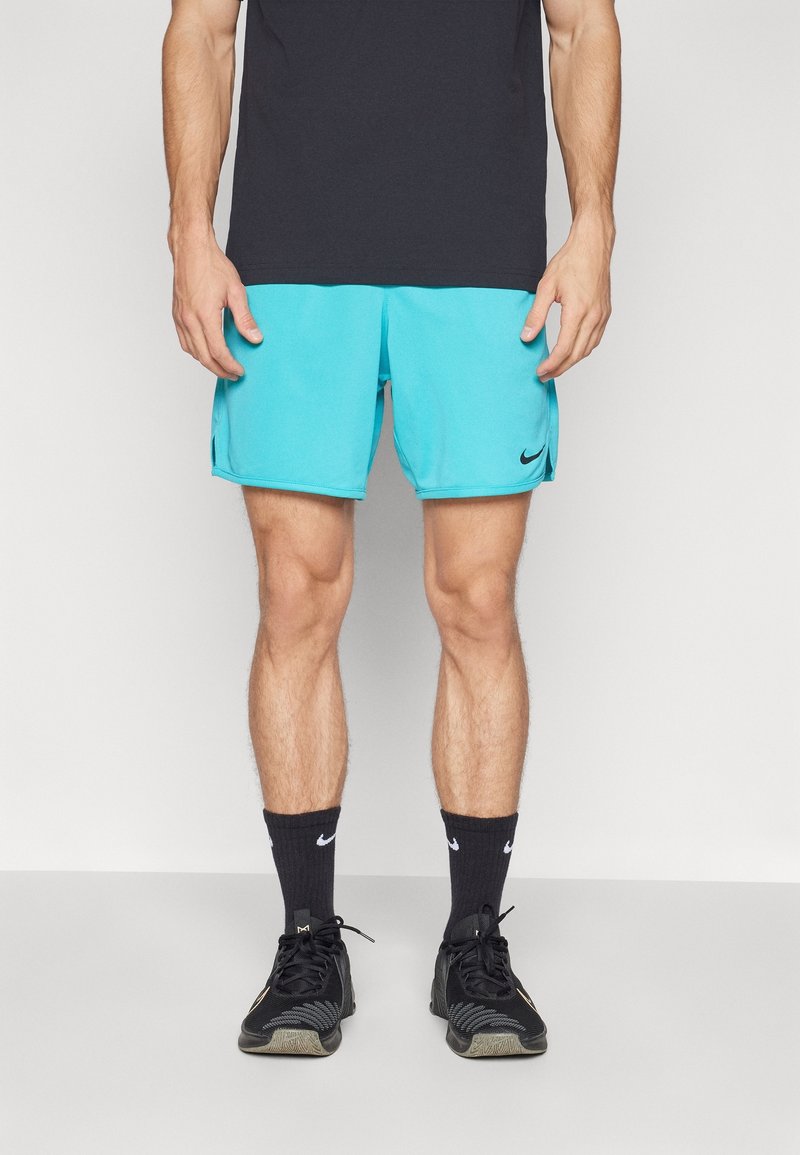 Personne portant un short de sport Nike turquoise, des chaussettes Nike noires, des chaussures de sport noires et un t-shirt noir, debout devant un fond uni.