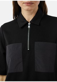 Zwarte polo met een ritssluiting bij de kraag. Bevat twee grote donkere stoffen zakken. Materiaal lijkt glad met een lichte glans.