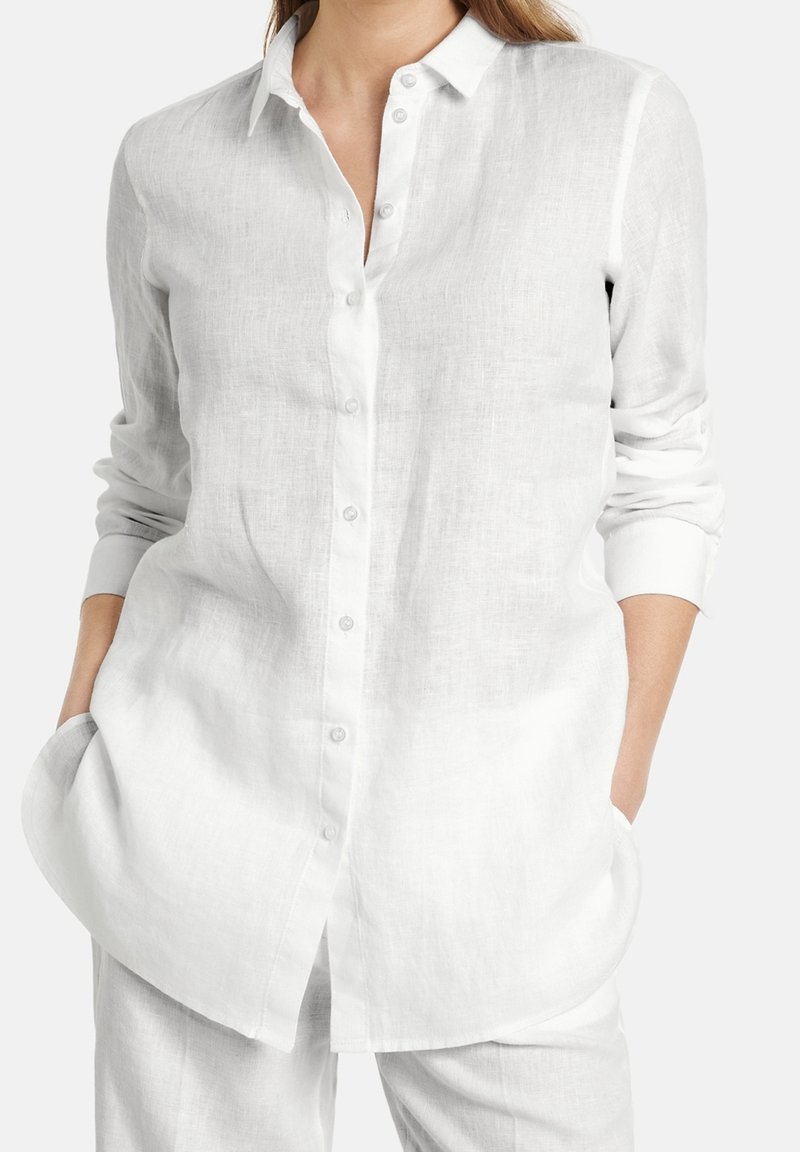 Femme portant une chemise blanche en lin à manches longues avec col et pantalon blanc en lin, mains dans les poches, sur fond uni.