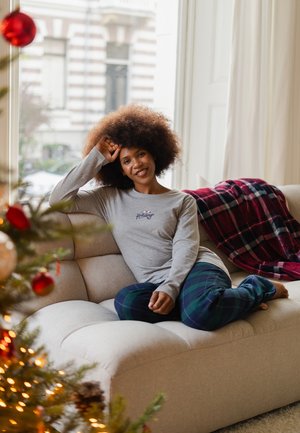 Chemise gris à manches longues avec le texte "Holiday", associée à un pantalon de pyjama à carreaux bleu foncé. Assis sur un canapé beige clair avec un jeté à carreaux rouges à proximité.