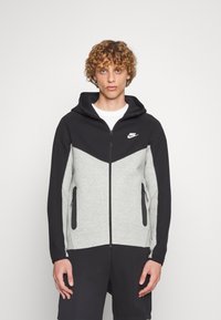 Nike zip-up hoodie med grå krop og sorte ærmer og hætte, tekstureret stof, to lynlåslommer i siderne og logo på brystet.