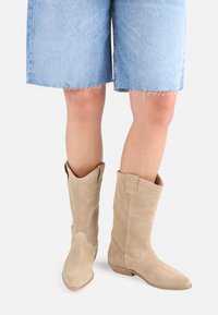 Bottes de cow-boy en suède beige avec des pointes fines, hauteur mi-mollet et détails de couture décoratifs. Portées avec un short en jean.