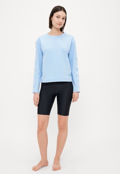 Light blue sweatshirt with long sleeves, features white logo print on sleeves, paired with black fitted shorts, barefoot. Simple design, casual style.