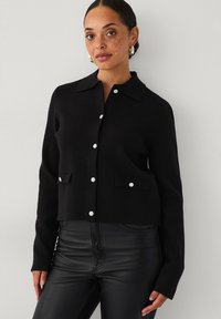 Zwarte cropped cardigan met een klassieke kraag, voorzien van knoopsluiting en twee buitenzakken. De stof lijkt zacht met een textuurafwerking.