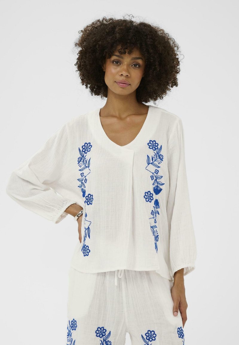 Femme aux cheveux bouclés portant un chemisier blanc ample et un pantalon avec une broderie florale bleue, debout avec une main sur la hanche.