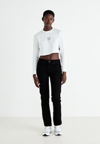 Fehér, rövidített, hosszúujjú póló logóval, fekete, slim-fit farmernadrággal és szürke kiegészítőkkel ellátott fehér sportcipővel, sima háttérrel.