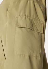 Khaki-farvet stoflomme med en rektangulær klap sikret med syninger på et beklædningsstykke, sandsynligvis bukser eller shorts.