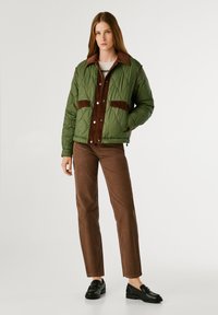 Giacca trapuntata verde oliva con parte frontale e tasche in corduroy marrone; abbinata a pantaloni in corduroy marrone e mocassini neri.