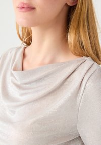 Jasnobeżowa bluzka z dekoltem w zakładkę, miękką teksturą i subtelnym połyskiem, z marszczeniem na ramieniu.