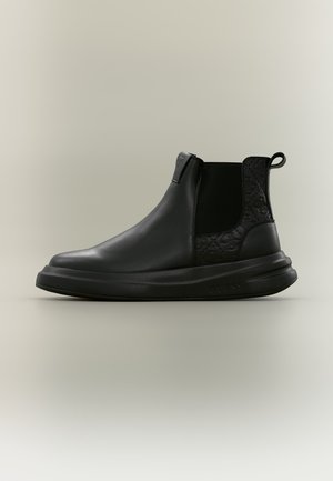Bottine noire en cuir à enfiler avec semelle épaisse, panneaux latéraux élastiques, motif floral embossé sur le talon et languette à l'arrière.