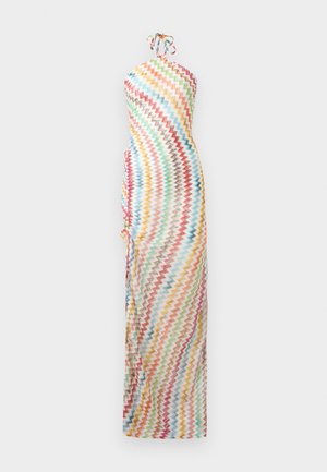 Femme portant une robe maxi dos nu à motif zigzag multicolore, debout les mains derrière le dos sur un fond uni.