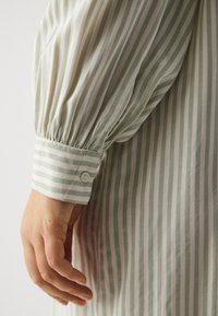 Chemise rayée présentant un schéma de couleurs vert clair et blanc, avec un poignet boutonné et un détail froncé à la manche. Tissu doux et léger.