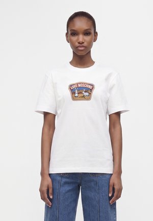 Νεαρή γυναίκα που φοράει λευκό t-shirt Love Moschino με σχέδιο λαγού και σκύλου και μπλε τζιν, στέκεται μπροστά σε απλό φόντο.