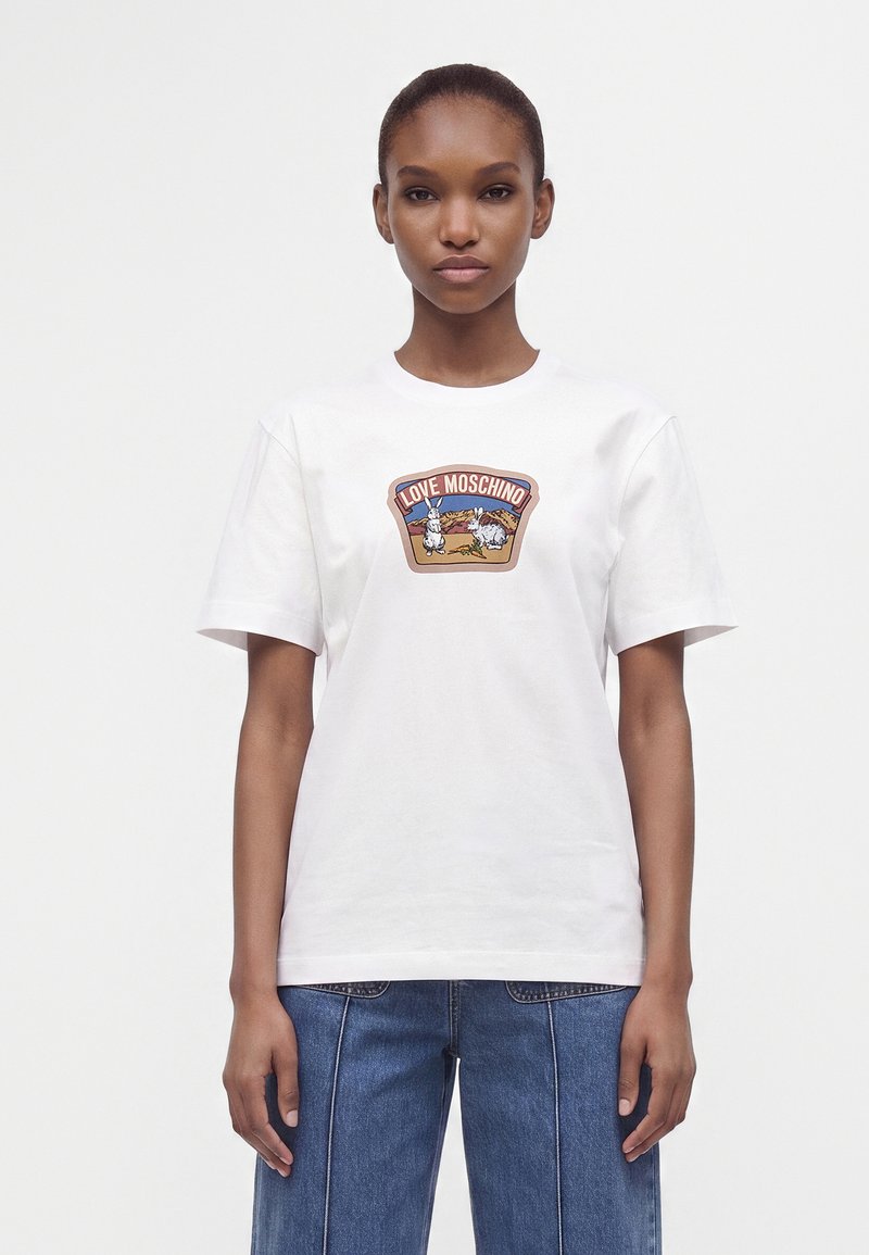 Νεαρή γυναίκα που φοράει λευκό t-shirt Love Moschino με σχέδιο λαγού και σκύλου και μπλε τζιν, στέκεται μπροστά σε απλό φόντο.