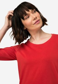Maglietta rossa a maniche lunghe con scollo rotondo e superficie liscia, caratterizzata da una silhouette aderente e cuciture sulle spalle discrete.