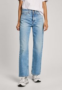 Jeans de tiro alto en azul claro con un diseño de pierna recta, cierre de botón visible y un ligero desvanecimiento. Combinados con zapatillas blancas.