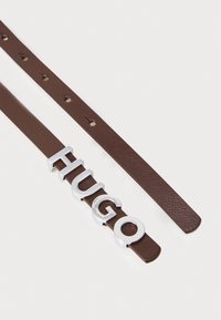 Brauner Ledergürtel mit einem auffälligen silbernen "HUGO"-Logo. Der Riemen hat mehrere Löcher zur Anpassung und eine strukturierte Oberfläche.