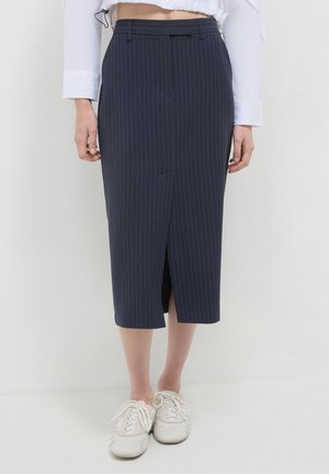 Femme portant une jupe midi bleu marine à rayures avec fente devant, associée à des baskets blanches à lacets et une chemise blanche à manches longues.