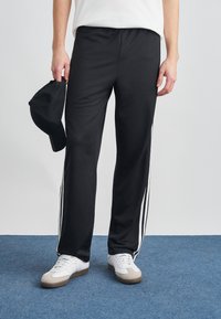 Μαύρο αθλητικό παντελόνι με υφή, με λευκές ρίγες στα πλάγια και άνετη γραμμή. Κρατιέται στο χέρι με μαύρο καπέλ ο.