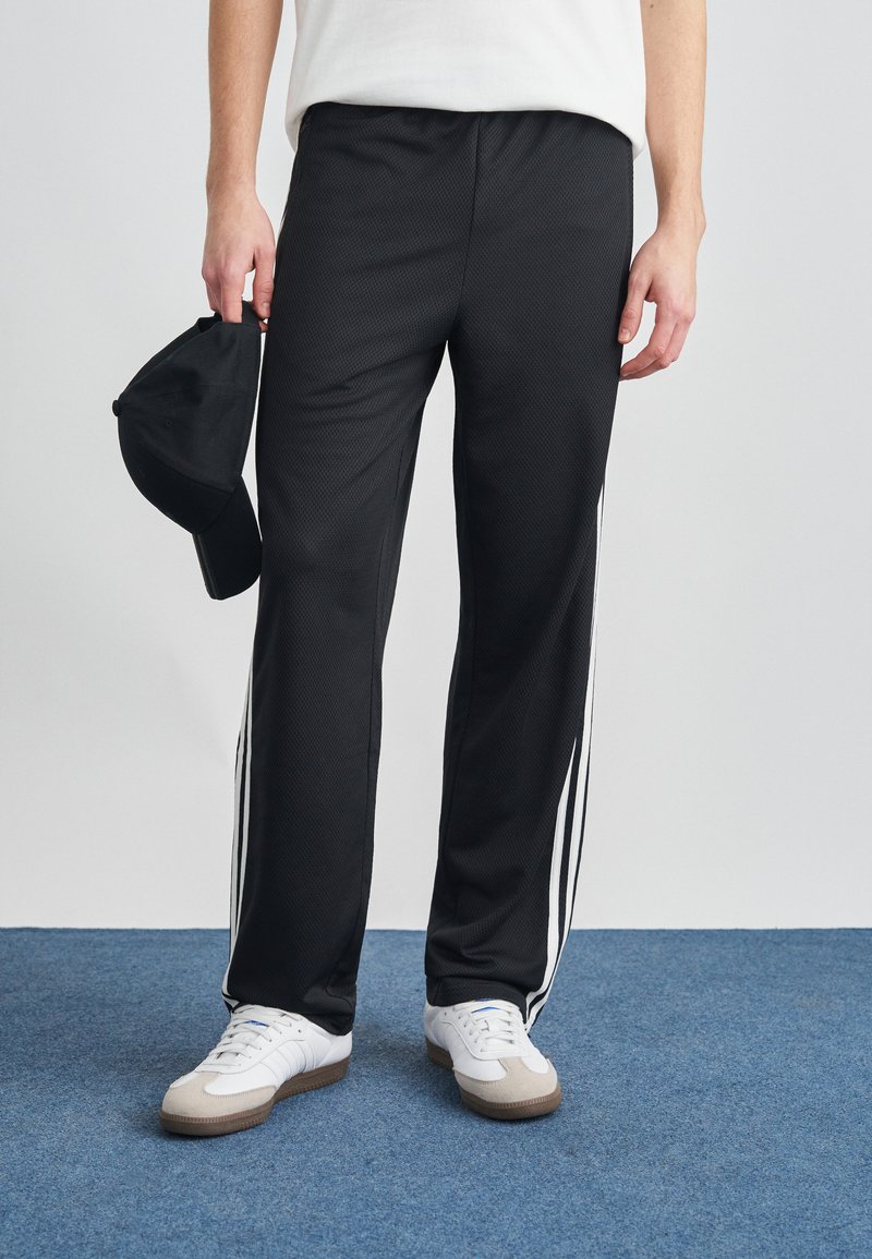 Μαύρο αθλητικό παντελόνι με υφή, με λευκές ρίγες στα πλάγια και άνετη γραμμή. Κρατιέται στο χέρι με μαύρο καπέλο.