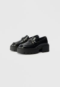 Black leather loafers with a chunky platform, featuring a decorative strap and metal hardware. Smooth texture with a rounded toe design.