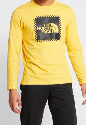 T-shirt à manches longues jaune en coton, avec un logo carré noir orné de bandes diagonales et de texte jaune, col rond.