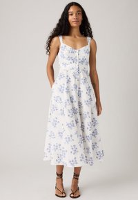 Donna che indossa un abito midi senza maniche bianco con stampa floreale blu e bottoni frontali, abbinato a sandali neri con cinturini, in piedi su uno sfondo neutro.