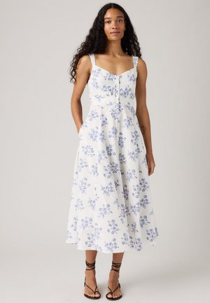 Femme portant une robe midi sans manches blanche avec un imprimé floral bleu et des boutons à l'avant, associée à des sandales noires à lanières, se tenant devant un fond uni.