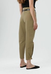 Kaki taps toelopende broek met hoge taille, voorzien van een zwarte riem, omslagen bij de zoom en een gladde textuur, gecombineerd met zwarte instapschoenen.