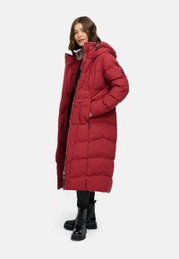 Cappotto puffer rosso lungo con cappuccio, caratterizzato da un design trapuntato, bottoni frontali e tasche laterali. Abbinato a stivali da combattimento neri e pantaloni.