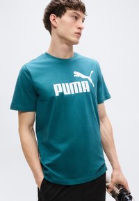 Τυρκουάζ βαμβακερό t-shirt με στρογγυλή λαιμόκοψη, που φέρει μεγάλο λευκό λογότυπο "PUMA" και μια εικόνα ενός αλματωδώς πούμα στο μπροστινό μέρος.