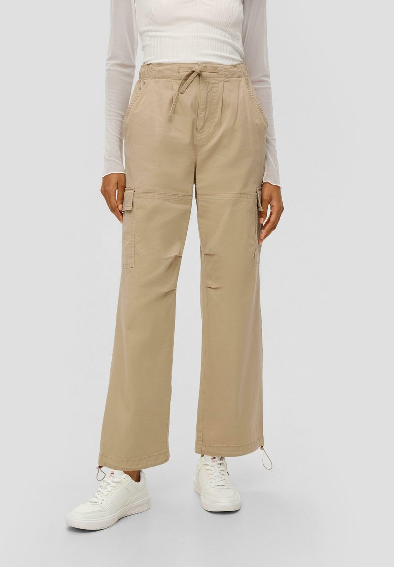 Beige cargobroek van katoen met een relaxte pasvorm, voorzien van zijzakken en verstelbare enkelbinders, gedragen met witte sneakers.