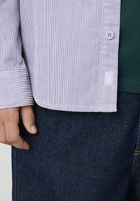 Chemise en velours côtelé violet clair avec des rayures verticales, fermeture par bouton et une étiquette sur l'ourlet, associée à un jean en denim bleu foncé.