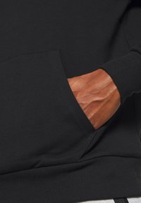 Schwarzer Hoodie aus weichem Material mit einer Kängurutasche. Die Bündchen und der Saum haben eine gerippte Textur. Sichtbar ist eine Hand, die in der Tasche ruht.