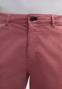 Pantaloni di cotone rosa con una texture liscia, dotati di chiusura a bottone e dettagli cuciti lungo la parte frontale e le tasche.