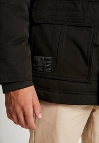 Černá all-weather bunda s hladkou texturou, obsahující nášivku, kapsy a přiléhavý manžet. Ruka umístěná na boku.