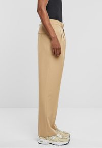 Beige maßgeschneiderte Hosen aus einem glatten Stoff, mit einem gefalteten Design und geradem Schnitt, getragen mit hellen Sneakers.