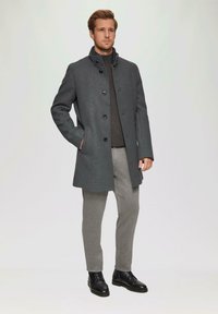 Donkergrijze wollen jas met een hoge kraag, knopen aan de voorkant en zijzakken, gecombineerd met grijze broek en zwarte leren schoenen.