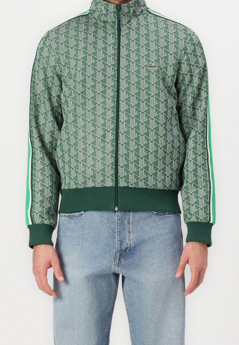 Personne portant une veste zippée verte à motifs avec des poignets et un ourlet côtelés, associée à un jean bleu clair.