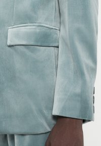 Svijetloplavi baršunasti sako s reverima, bočnim džepom i tri tamna gumba na manžetama. Glatka tekstura s blagim sjajem.