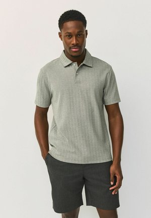 Mann trägt ein hellgraues kurzärmeliges Poloshirt und dunkelgraue Shorts, steht vor einem einfarbigen hellen Hintergrund.