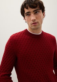 Roter strukturierter Strickpullover mit Rundhalsausschnitt, versehen mit einem Wabenmuster und gerippten Bündchen, vor neutralem Hintergrund.