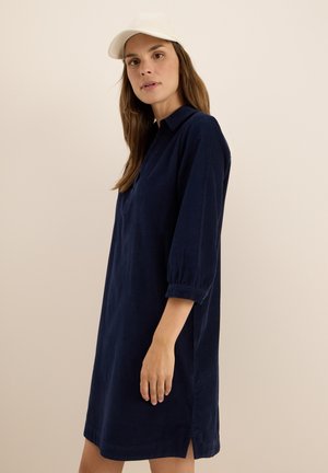 Donna con capelli lunghi che indossa un vestito blu navy a tre quarti e un cappellino da baseball beige, in piedi davanti a uno sfondo beige uniforme.