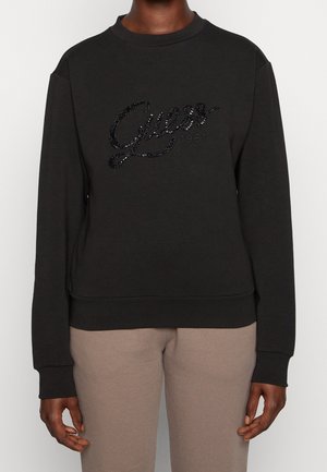 Black long-sleeve sweatshirt with "Guess 1981" in black sequins worn with beige pants, shown from neck to hips, front view.