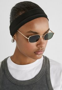 Occhiali da sole con una montatura dorata e lenti scure, indossati con una fascia per capelli in tessuto nero. L'individuo ha un outfit a strati grigio e bianco.