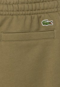 Pantalones deportivos de color verde aceituna, con textura suave, cinturilla elástica y un pequeño logo de cocodrilo bordado, con un bolsillo parche trasero.