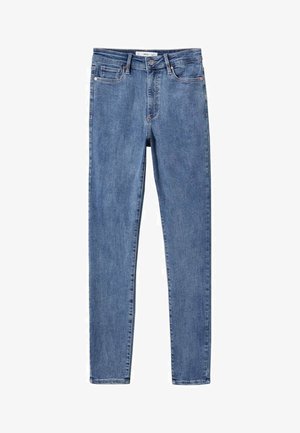 Person iført en sort blazer, grå sweater, blå skinny jeans og lyseblå spidse højhælede sko, der står på et neutralt gulv.